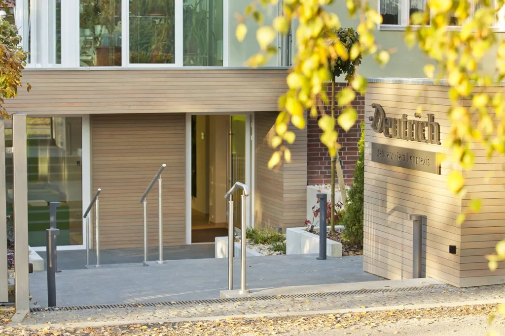 Ein einladendes Bild des Eingangsbereiches der Familienpraxis Deutrich bei spätmittaglichem Sonnenschein. Es führen zwei kleine Treppen bzw. Rampen hinunter zur Eingangstür. Falls Sie Fragen zur Zugänglichkeit haben, rufen Sie uns gerne an: 033397-6110