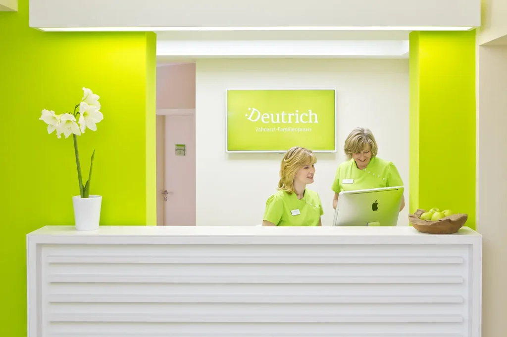 Der Empfangstresen der Praxis ist mit einer weißen Lilie dekoriert. Dahinter sitzen die Praxismanagerin Maria Stauch und Claudia Mroos aus der Verwaltung. Im Hintergrund sind Teile der Praxiswände in einem kräftigen Grün gestrichen.